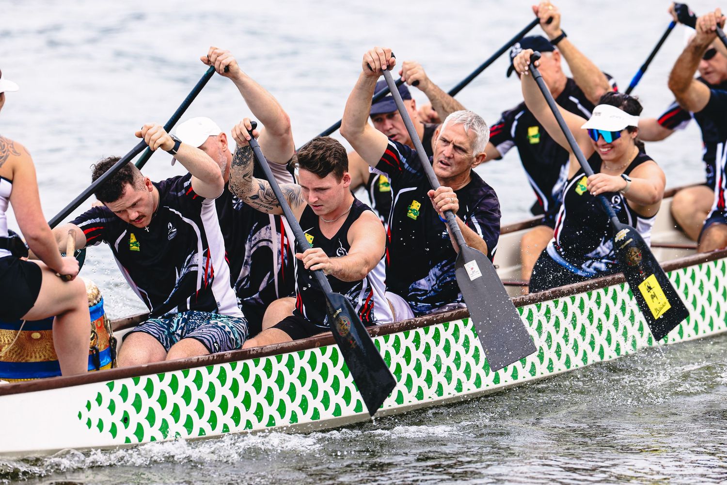 Dragon boat racers paddle in sync during a competitive race on the water while wearing black uniforms and life vests.