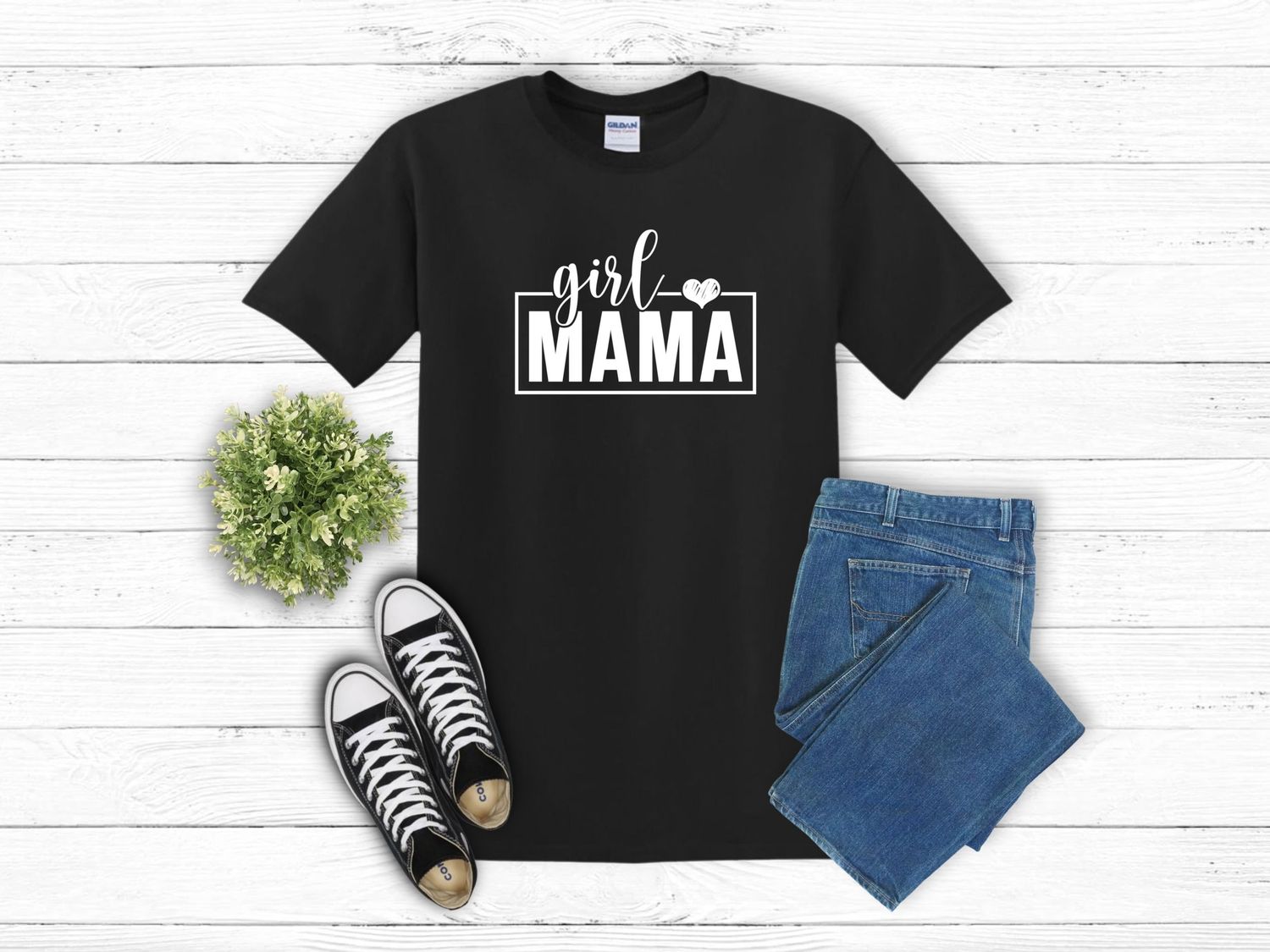 Flatlay outfit with black 'Girl Mama' t-shirt, blue jeans, classic black sneakers and small green plant decor on white wood.