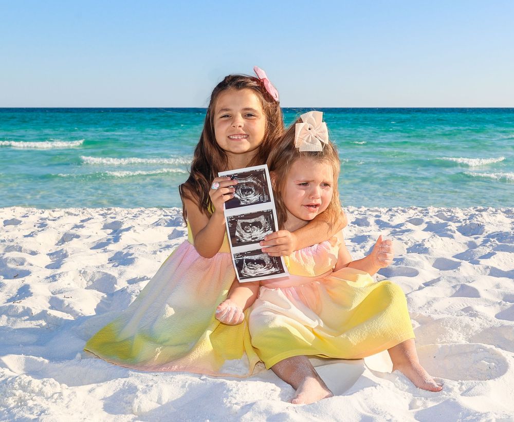 Two children in yellow dresses sit on a white sandy beach with turquoise ocean waves in the background.