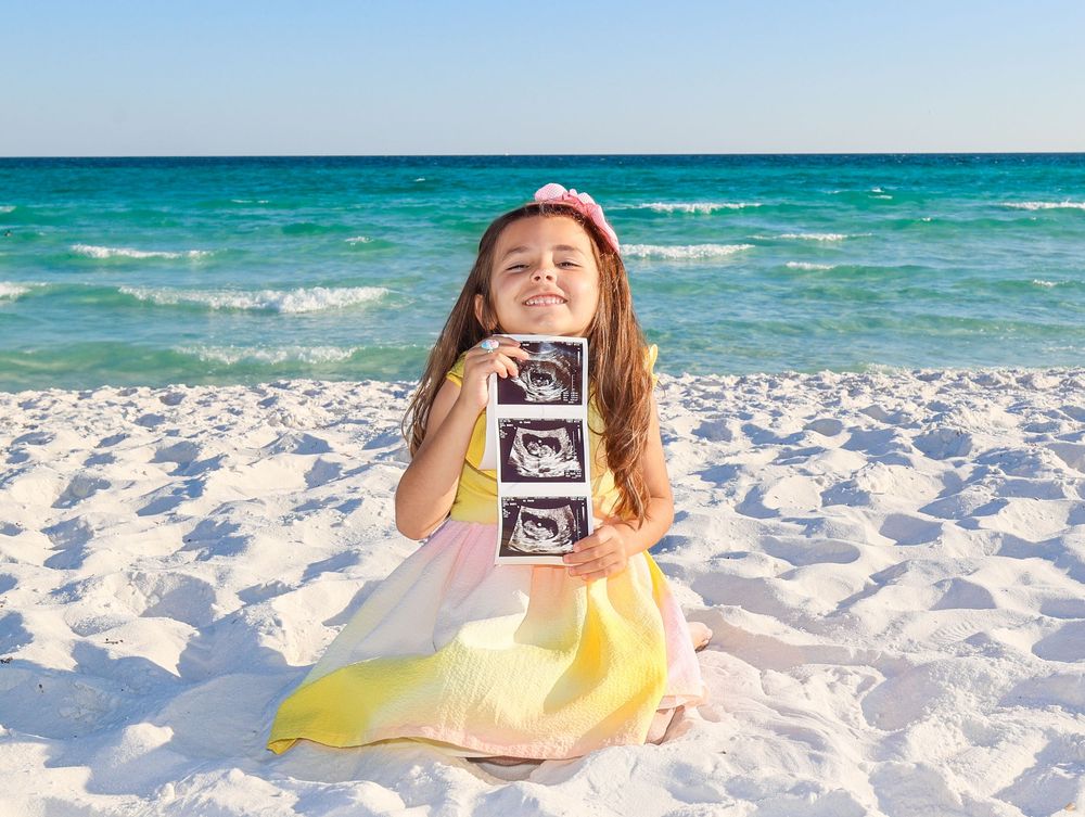 Someone in a yellow dress sits on a white sandy beach holding ultrasound photos with turquoise ocean waves behind them.