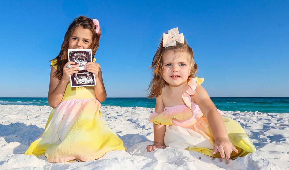 Two children in pastel dresses sit on white sand beach holding photographs while ocean waves roll in background.