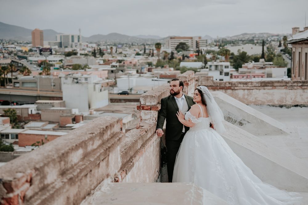 A couple in formal wedding attire pose together on a rooftop overlooking a cityscape with mountains in the distance.