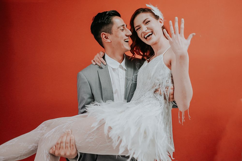 A joyful couple in wedding attire laugh together against a vibrant orange backdrop during their photoshoot.