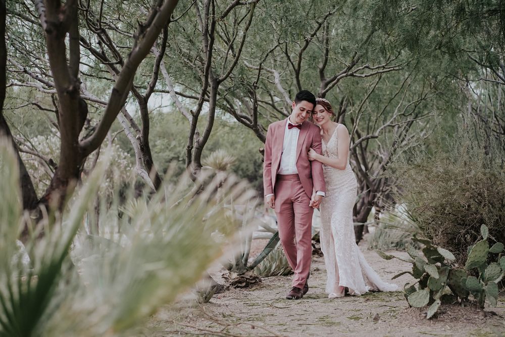 A dreamy desert wedding photo shows a couple embracing on a nature path surrounded by desert plants and trees.