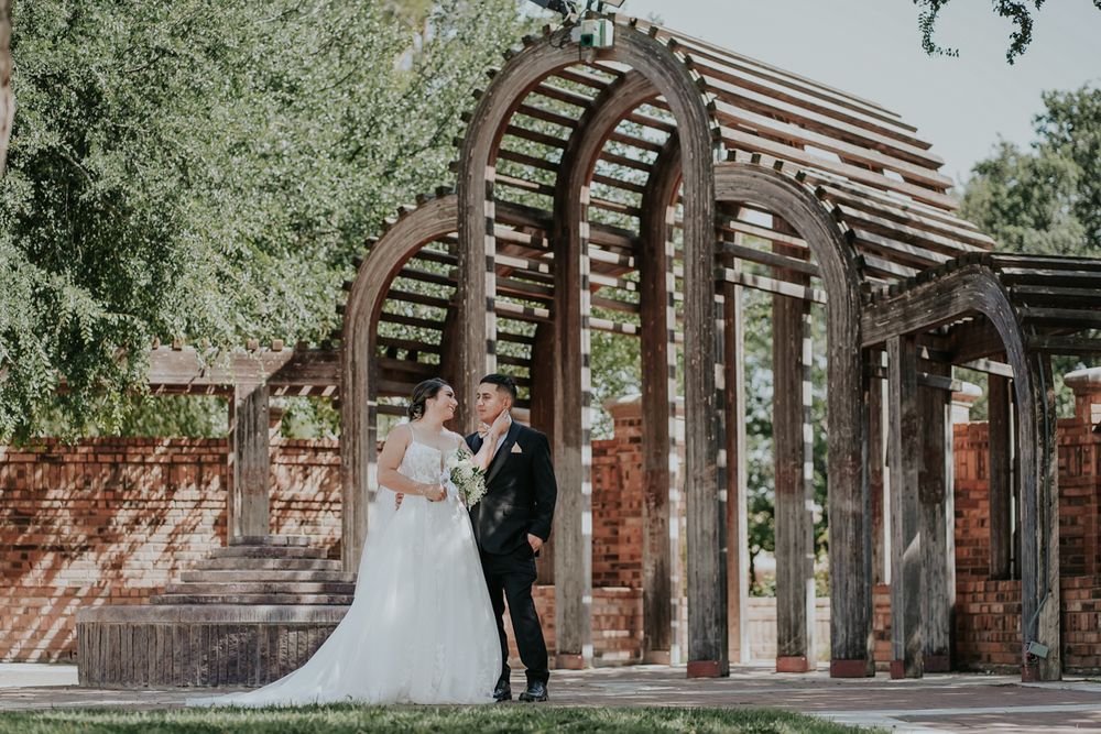 A couple shares a romantic moment under a wooden pergola in a lush garden setting.