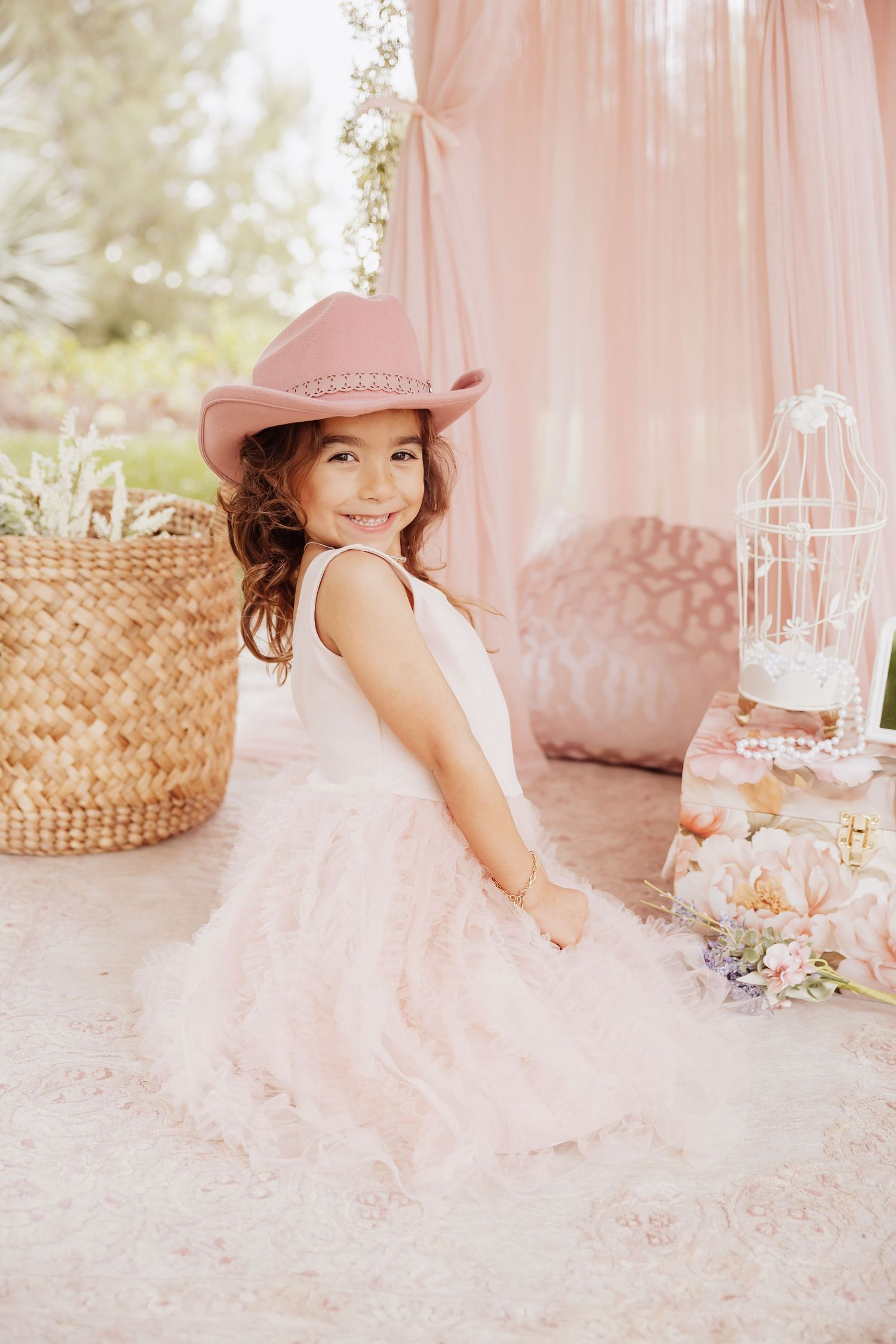Young girl in pink dress and cowgirl hat poses against dreamy backdrop with floral decor and wicker basket.
