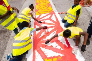 Workers in high-visibility vests paint a large red design on a concrete surface using stencils and rollers.