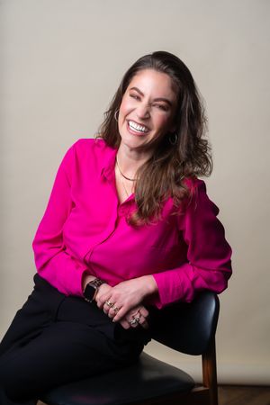 A person in a bright pink blazer and dark pants poses expressively while sitting in a wooden chair against a neutral wall.