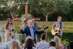 Outdoor worship service with musicians performing by a lake as people gather to listen in nature.
