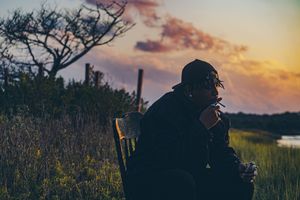 Silhouette of person photographing at sunset with dramatic orange and purple sky over grassy field.