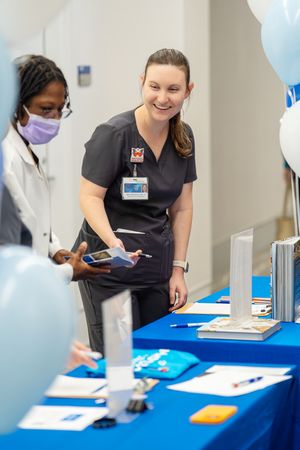 Healthcare professionals in scrubs interact at an event table decorated with blue balloons and informational materials.