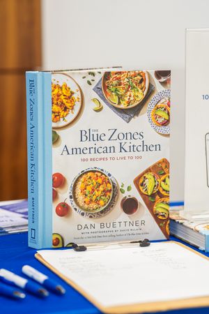 A cookbook titled 'Blue Zones American Kitchen' by Dan Buettner sits on a blue surface with pens and papers nearby.