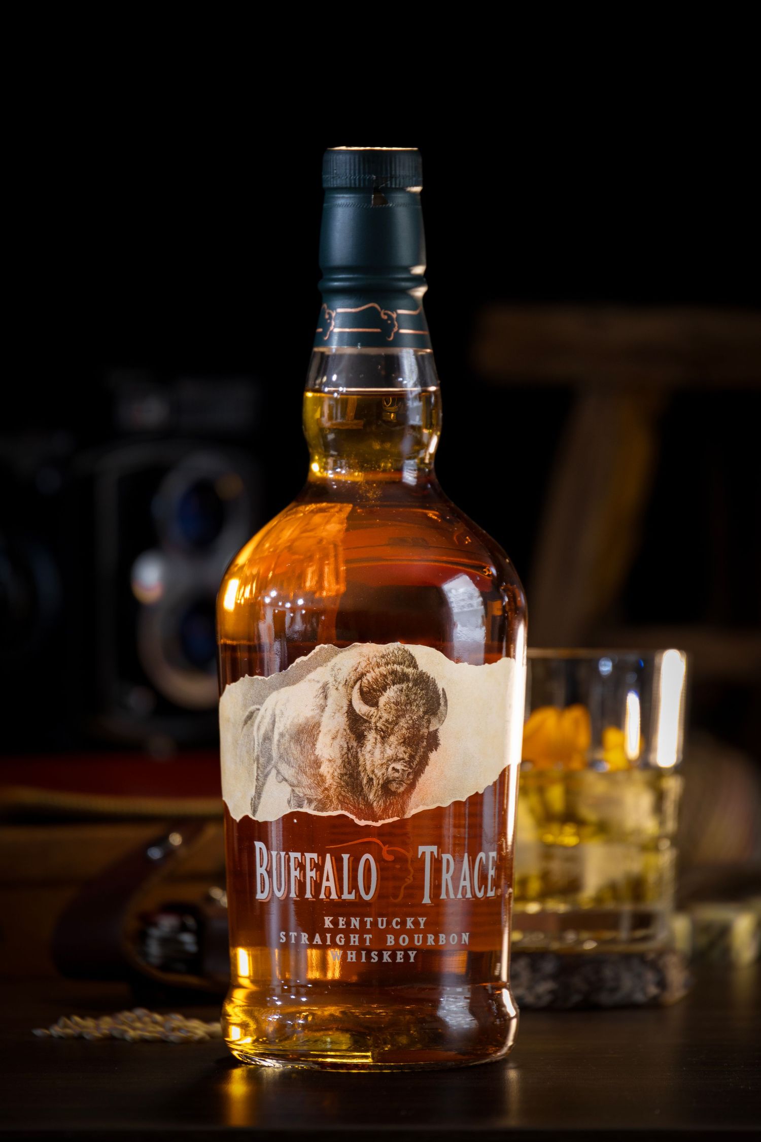 Buffalo Trace bourbon bottle beside a glass with ice and lemon peel, with a camera and wood background in soft lighting.