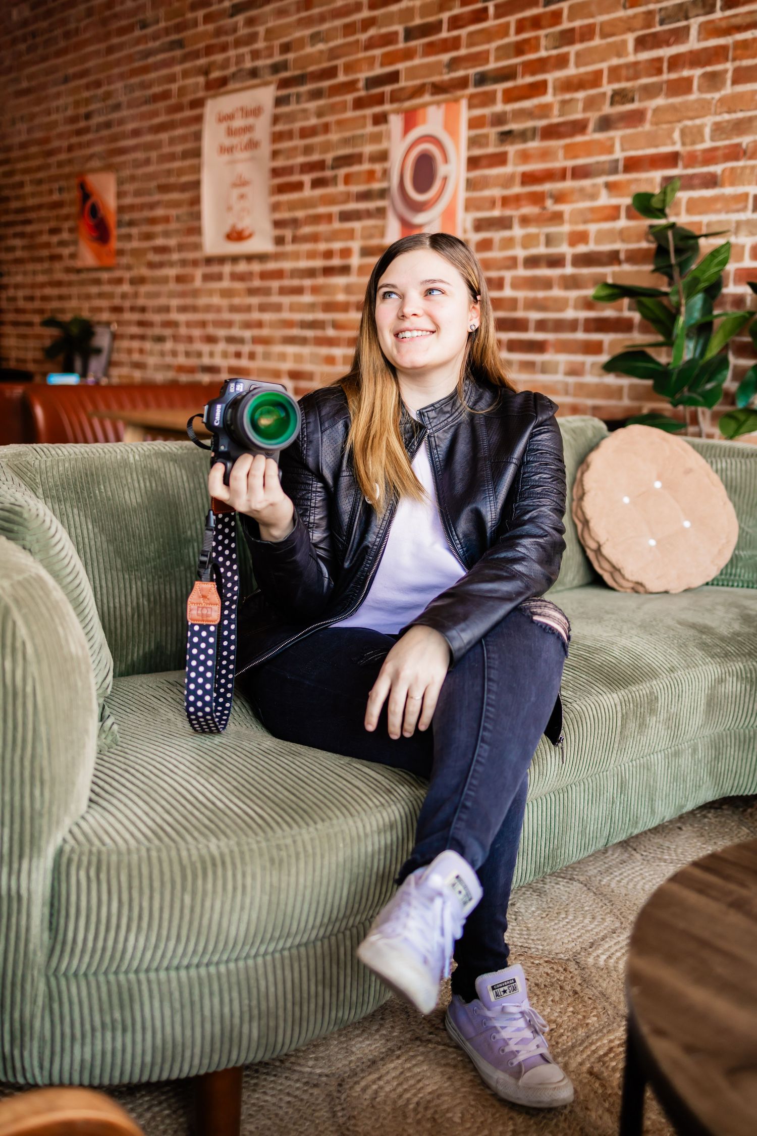 A photographer sits on a green couch holding professional camera gear in a room with exposed brick walls and modern decor.