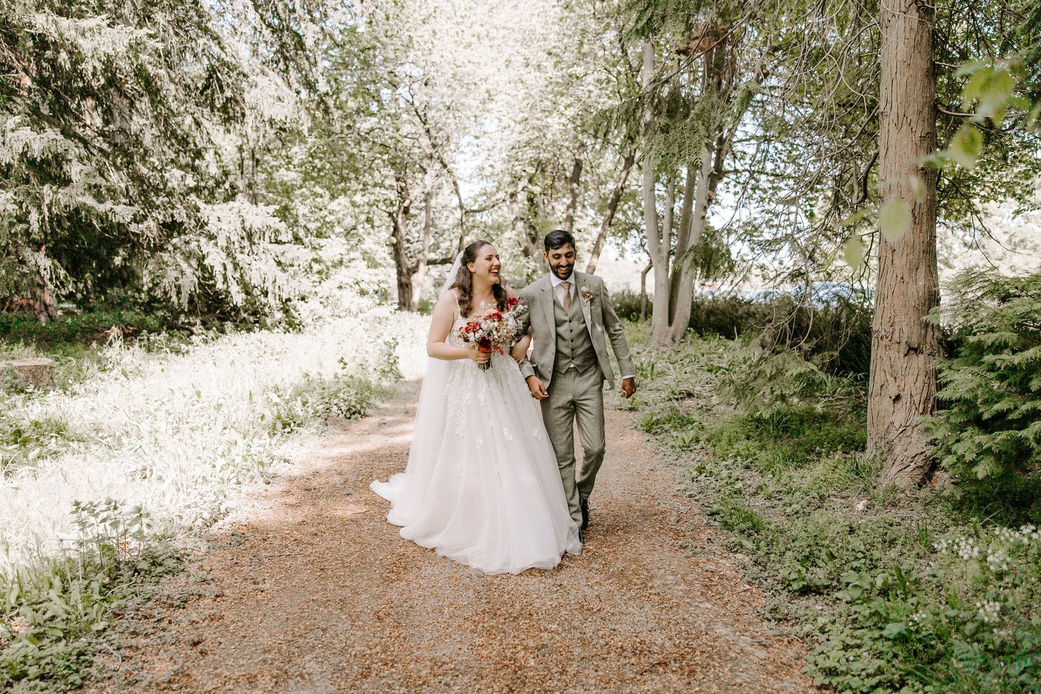 A couple walks hand in hand down a sunlit forest path, the bride in a flowing white dress and groom in a gray suit.