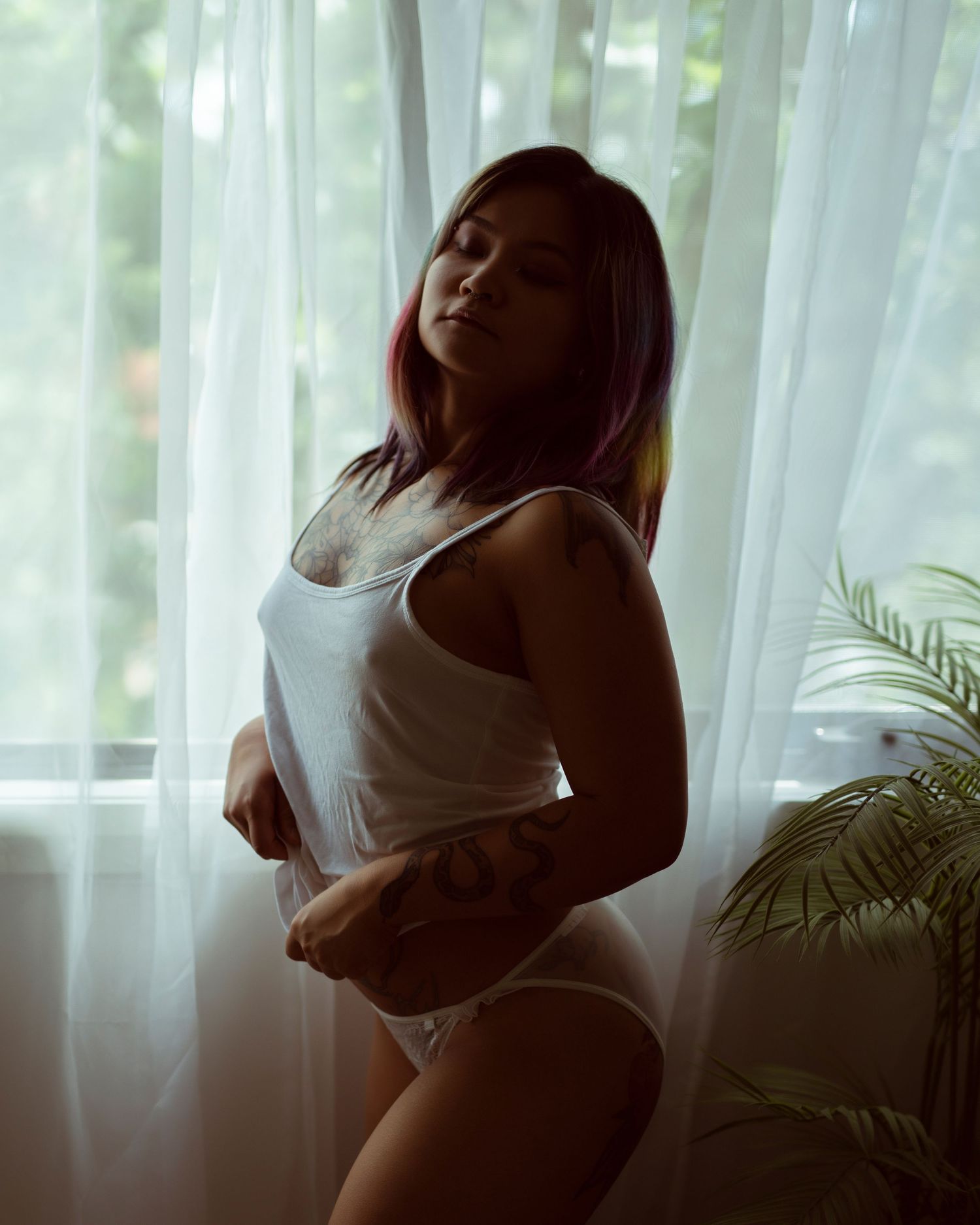 A silhouette figure wearing a loose white sweater sits on rumpled bedding against sunlit curtains in a moody boudoir scene.