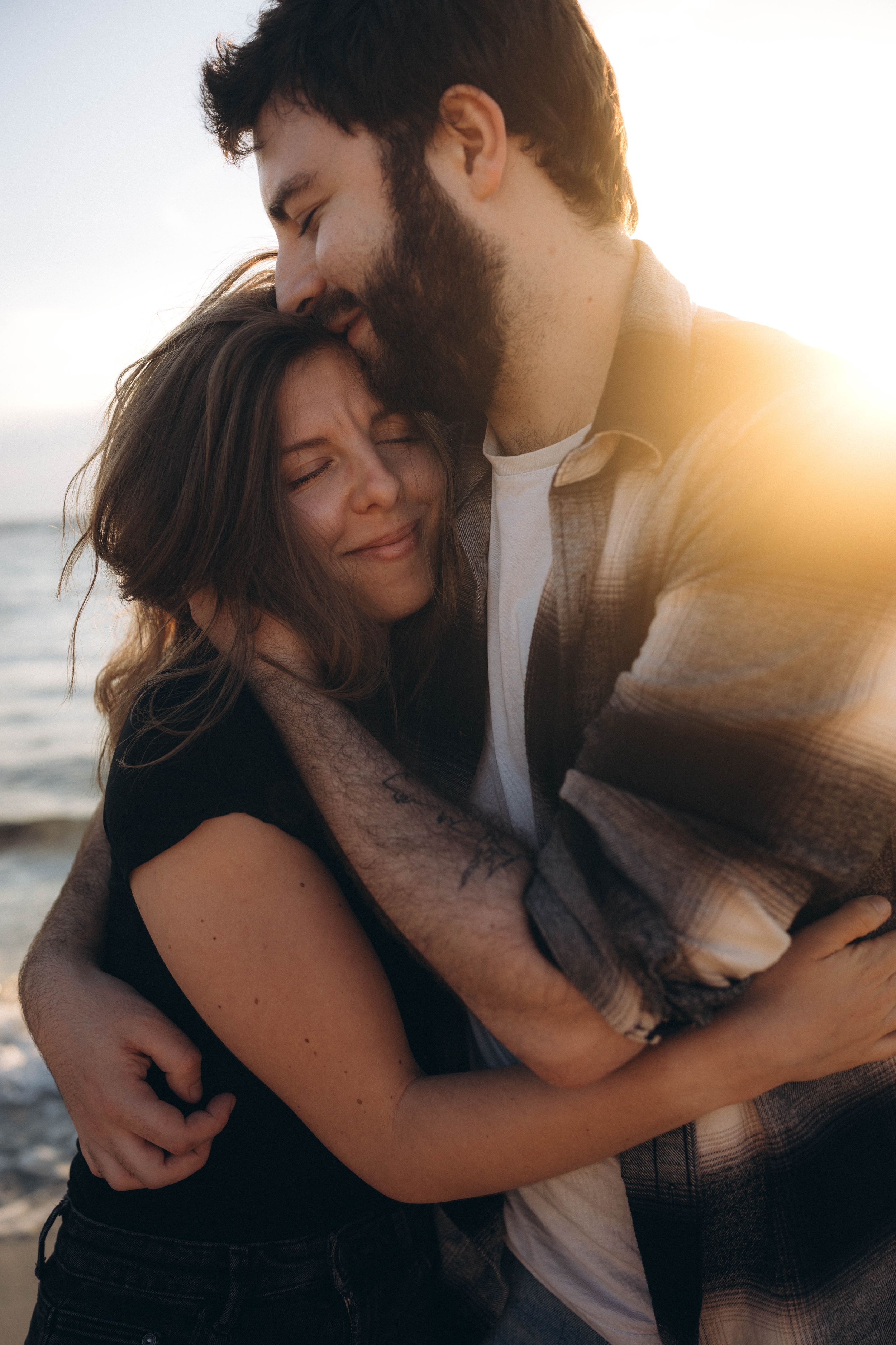 Romantic couple embracing at sunset on a beach, man kissing woman's head while she smiles with eyes closed.