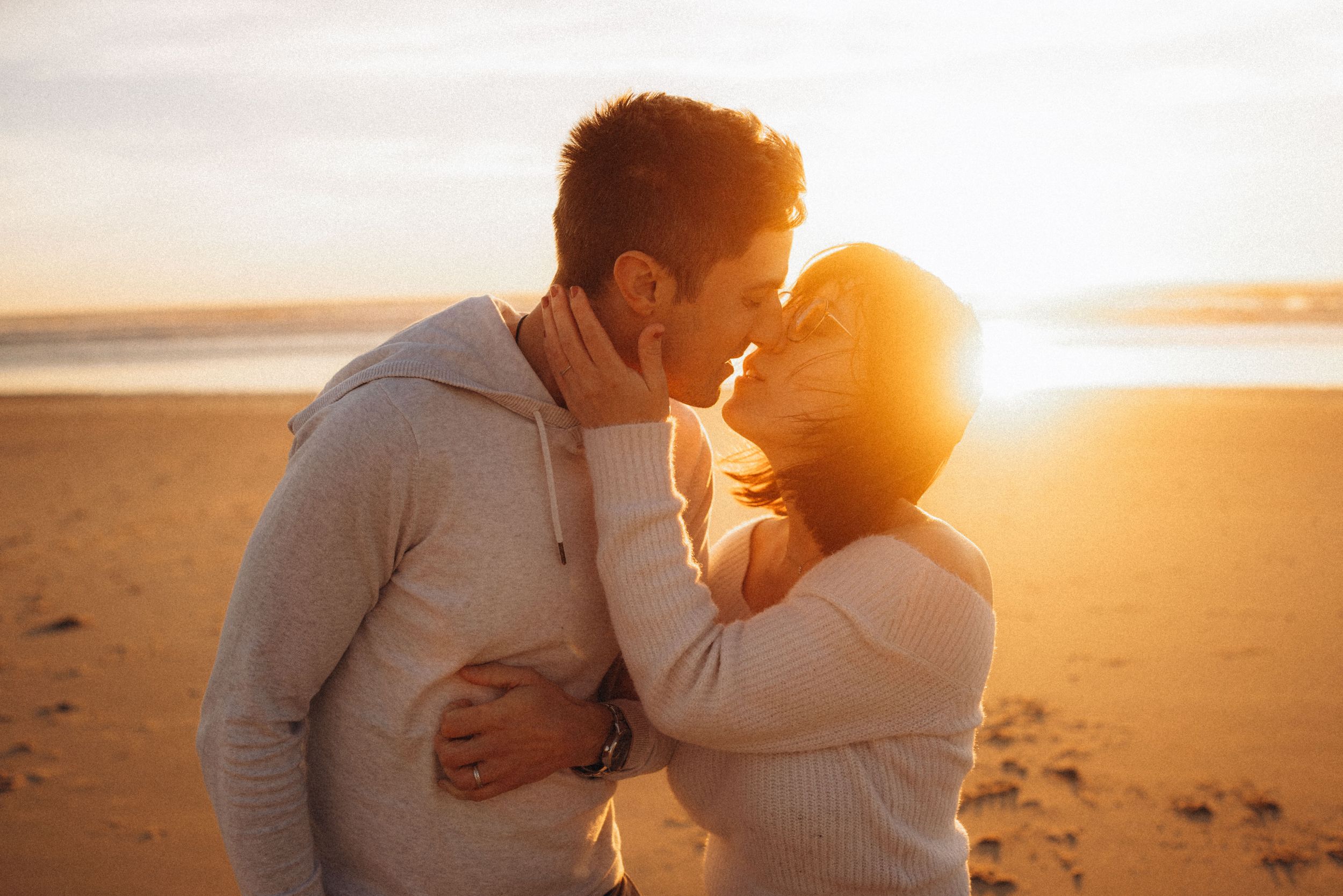 Romantic couple sharing a tender kiss on a sandy beach at golden sunset, embracing in warm sunlight.