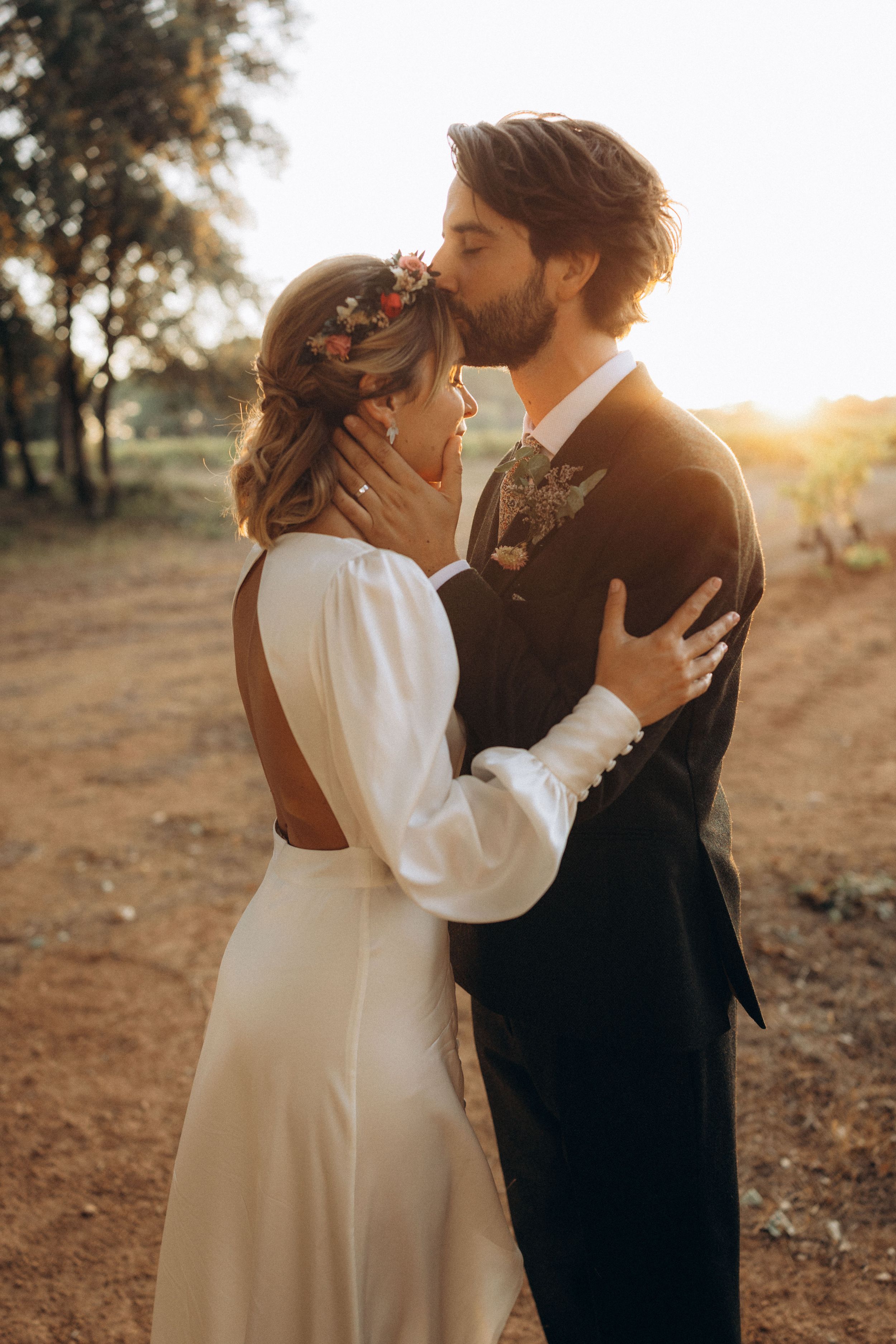 A bride and groom share a romantic kiss at golden hour, she wears a white backless gown with floral hair accessories.