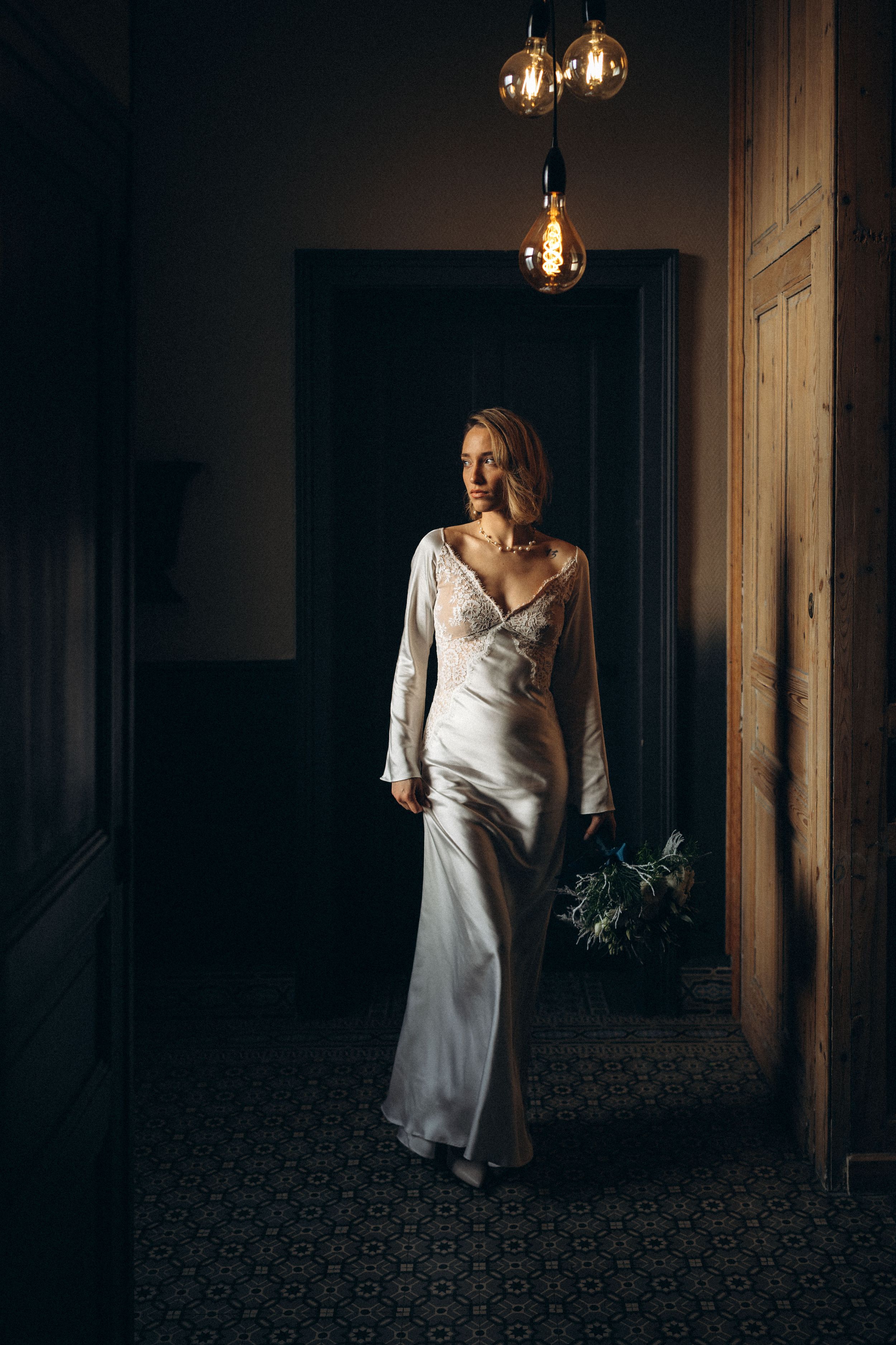 Elegant bride in a satin gown with lace sleeves holding a bouquet, walking through a dimly lit hallway.