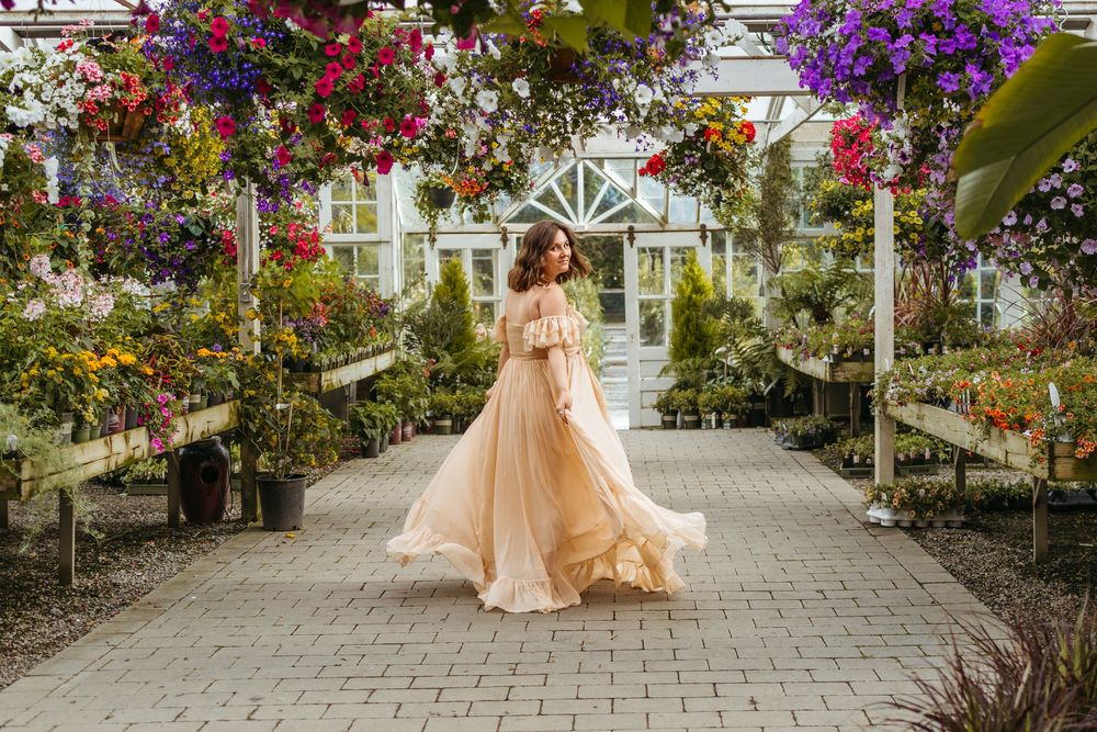 A peach ballgown dress twirls beneath vibrant hanging flowers in a greenhouse garden walkway.