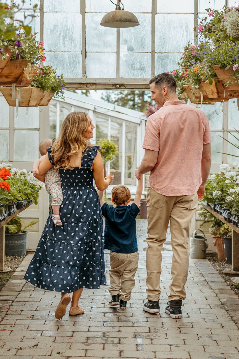 Family walking together through a sunny greenhouse filled with hanging flower baskets and potted plants.