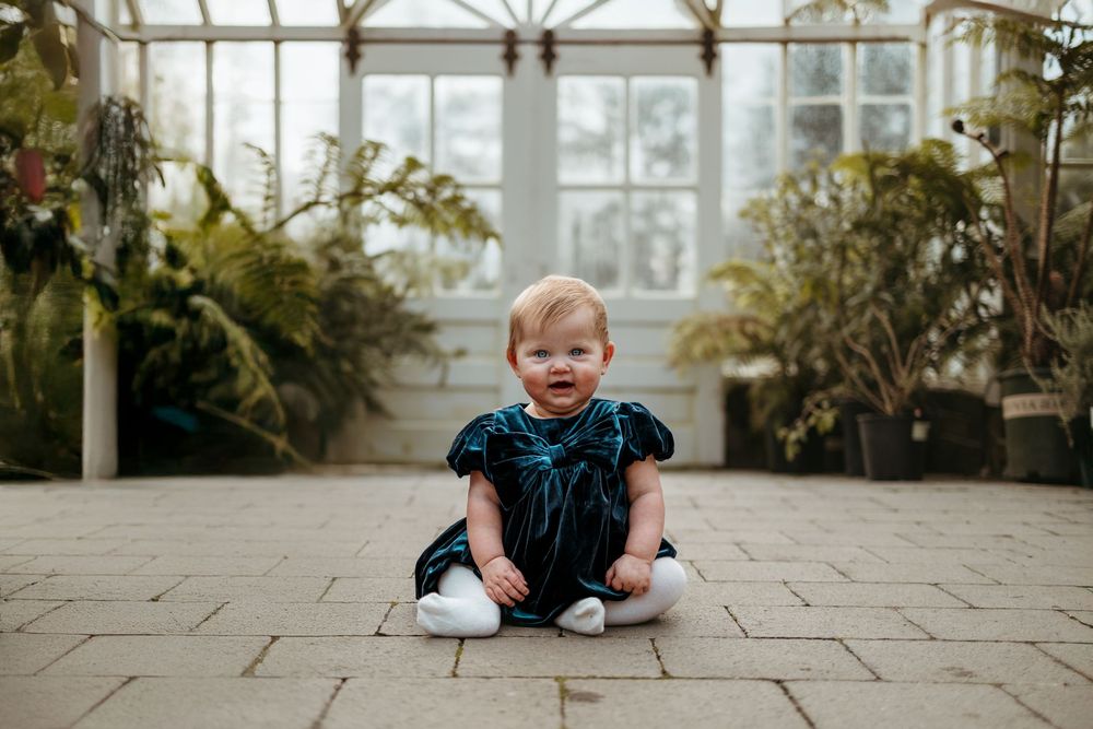Small child in velvet dress sits on stone floor inside bright conservatory filled with lush greenery and natural light.