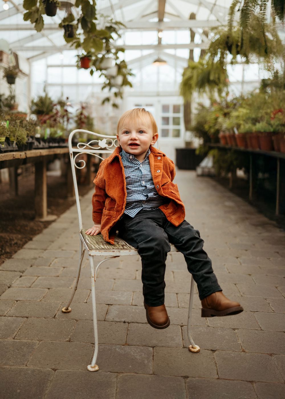 A small child sits on a white metal chair inside a bright greenhouse with hanging plants and stone flooring.