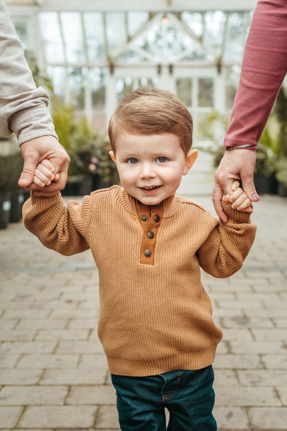 Close-up sequence of a toddler in brown sweater holding hands with adults while walking on stone pavement.