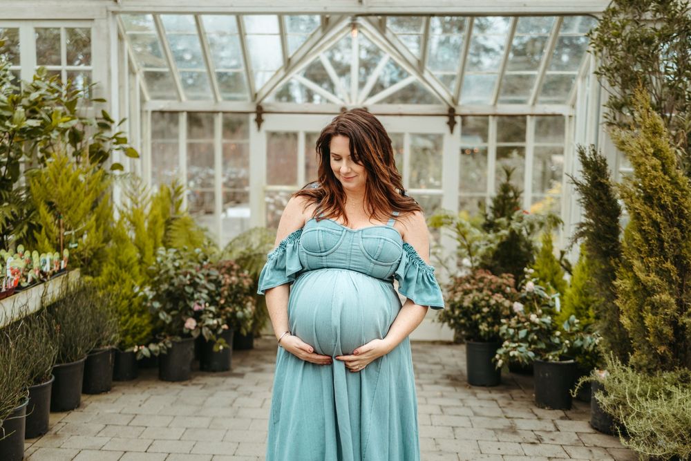 A pregnant woman in a turquoise off-shoulder dress stands in front of a greenhouse surrounded by potted plants.