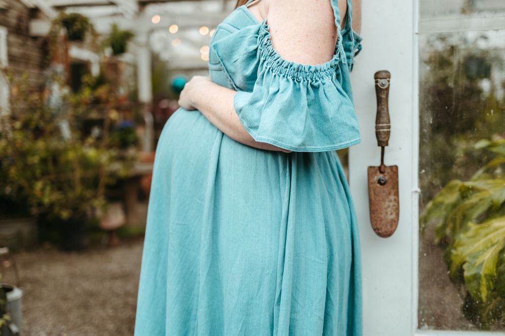 A pregnant figure in a flowing turquoise dress stands in a garden setting with a rustic shovel visible.