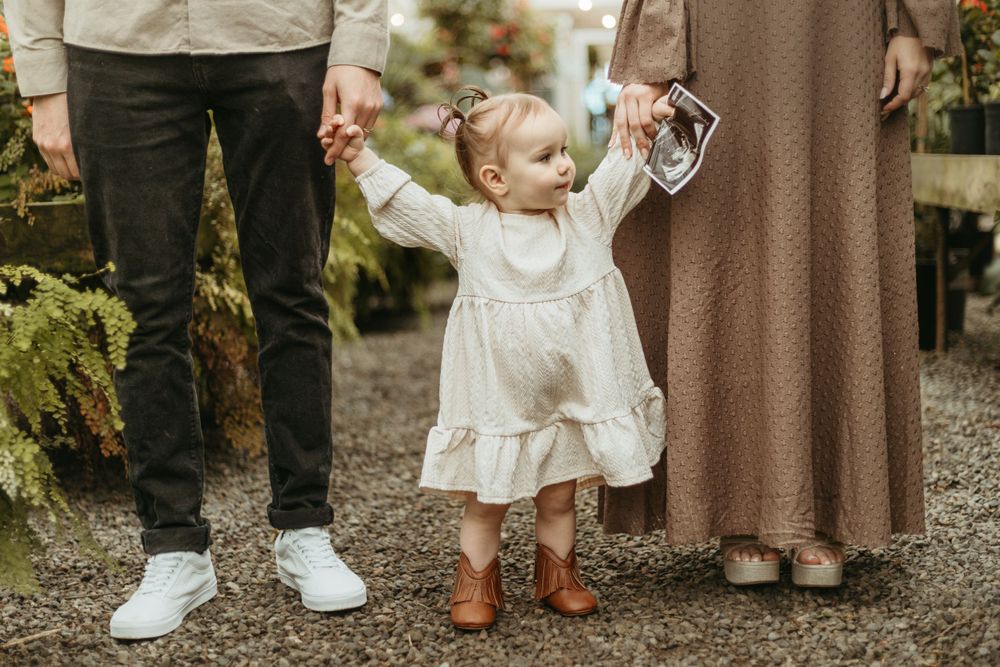 Toddler in white dress walking between parents in garden setting.