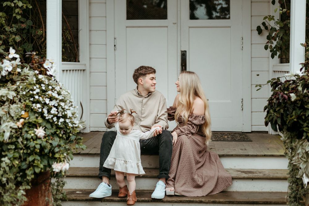 A family sits together on white porch steps surrounded by climbing vines and flowers.