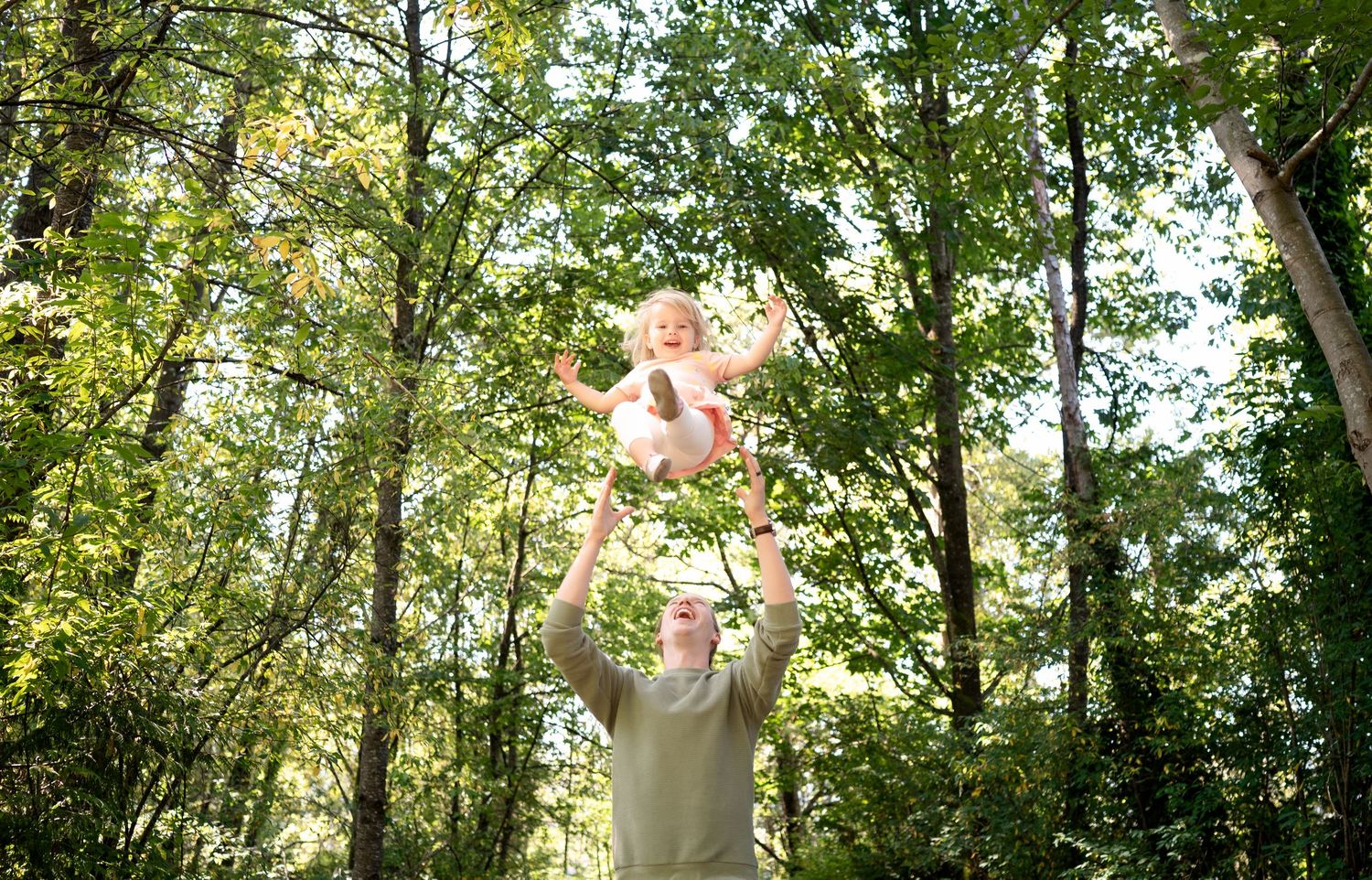 A figure lifts a baby high into the air against a sunlit forest backdrop of green trees.