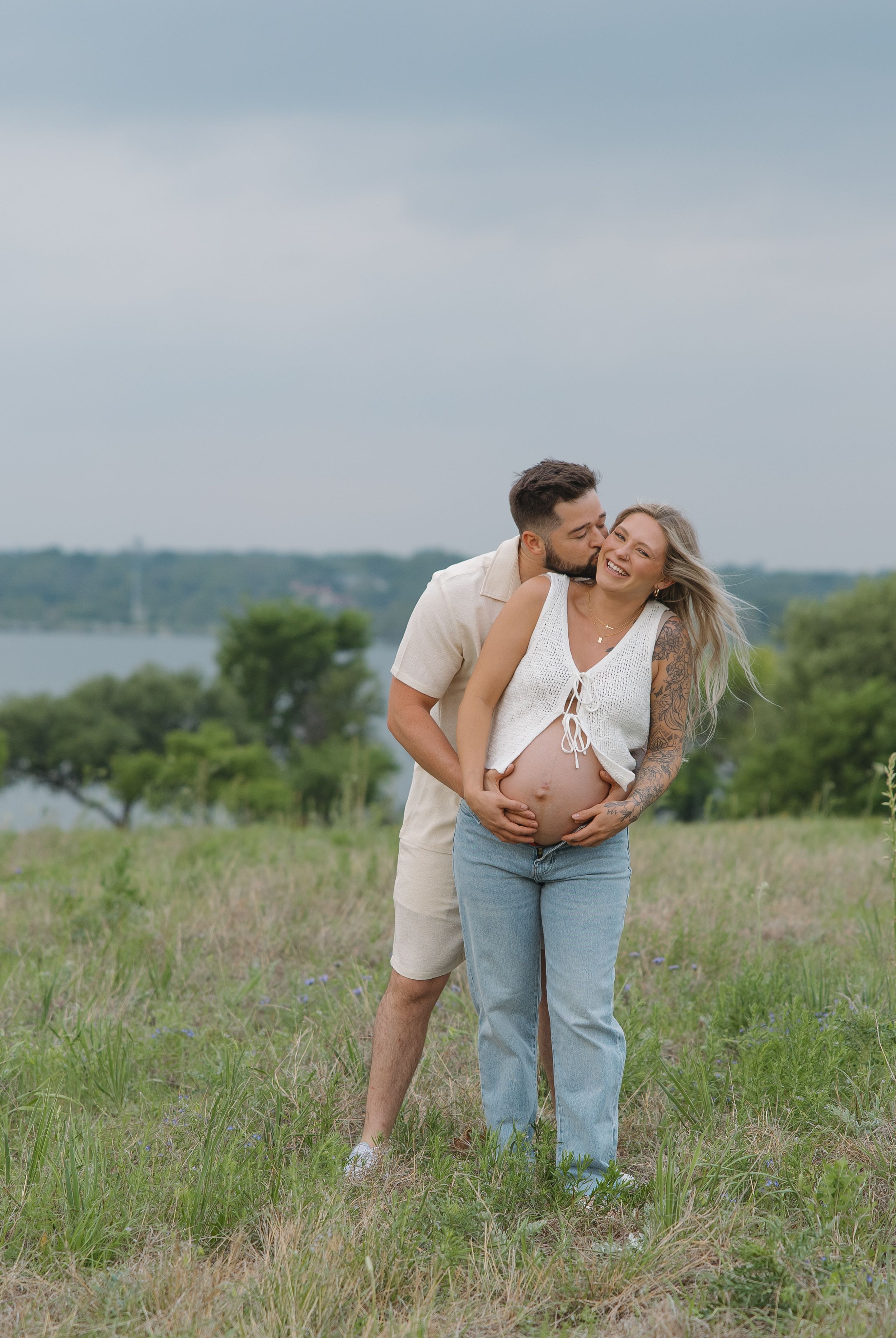 Expectant couple sharing intimate moment in grassy field with lake view and overcast sky.