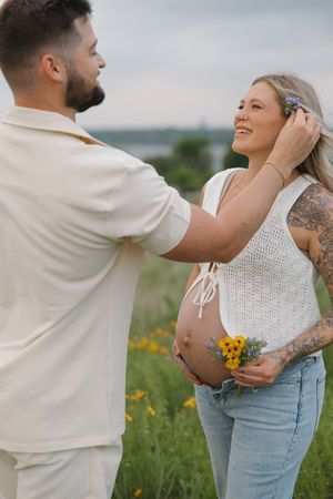 Expecting couple shares tender moment in field of wildflowers, wearing white and cream outfits at sunset.