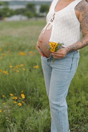 A pregnant belly in a white knit top with wildflowers held against it, standing in a meadow of yellow blooms.