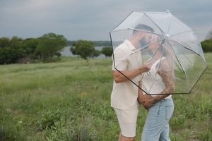 A couple shares a romantic moment under a clear umbrella while standing in a green field on an overcast day.