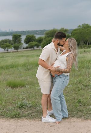 A couple shares an intimate moment on a hill overlooking a lake, surrounded by green trees and grass.