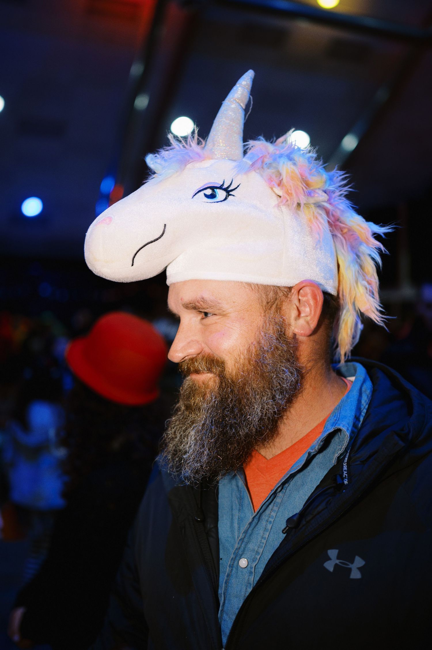 A bearded person wearing a whimsical white unicorn hat with colorful feathers and sparkly details at a nighttime event.