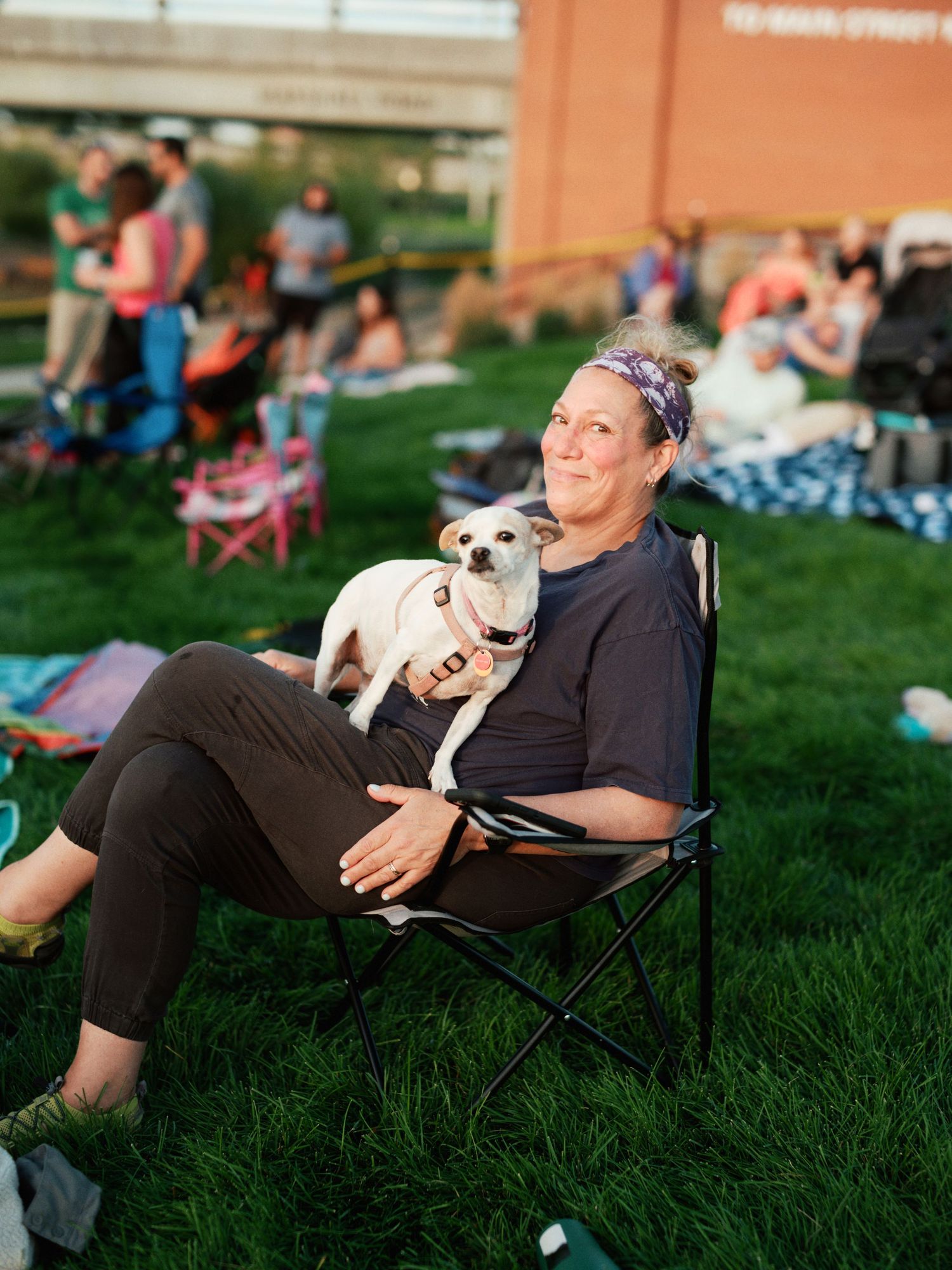A person enjoys an outdoor event on a camping chair with a small white dog sitting on their lap in a grassy area.