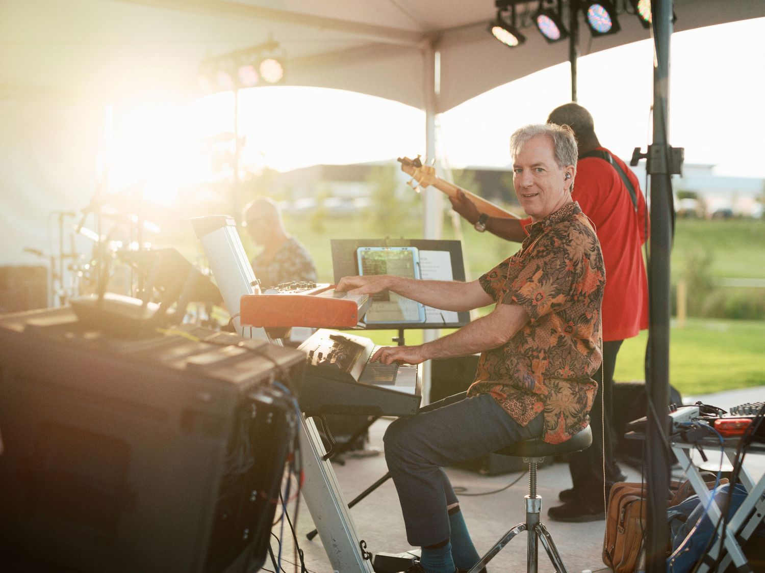 Musician playing keyboard on outdoor stage during golden hour sunset with warm sunlight glare in background.