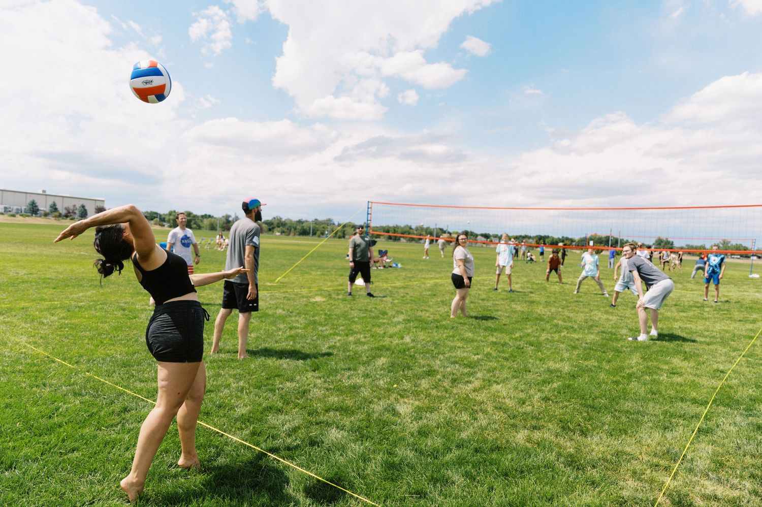 A group of people playing volleyball on a green field under a blue sky with white puffy clouds.