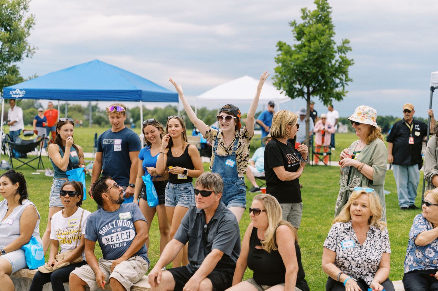 A crowd of festival-goers sitting on grass under a blue tent canopy at an outdoor summer event.