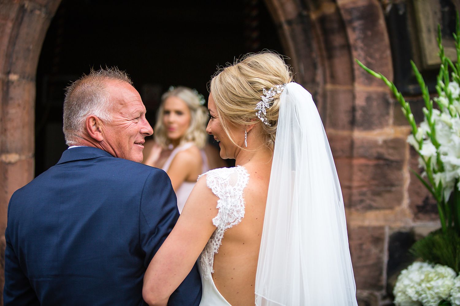 Father of the bride looks lovingly into his daughters eyes whilst being watched by a bridesmaid at a wedding in Cheshire