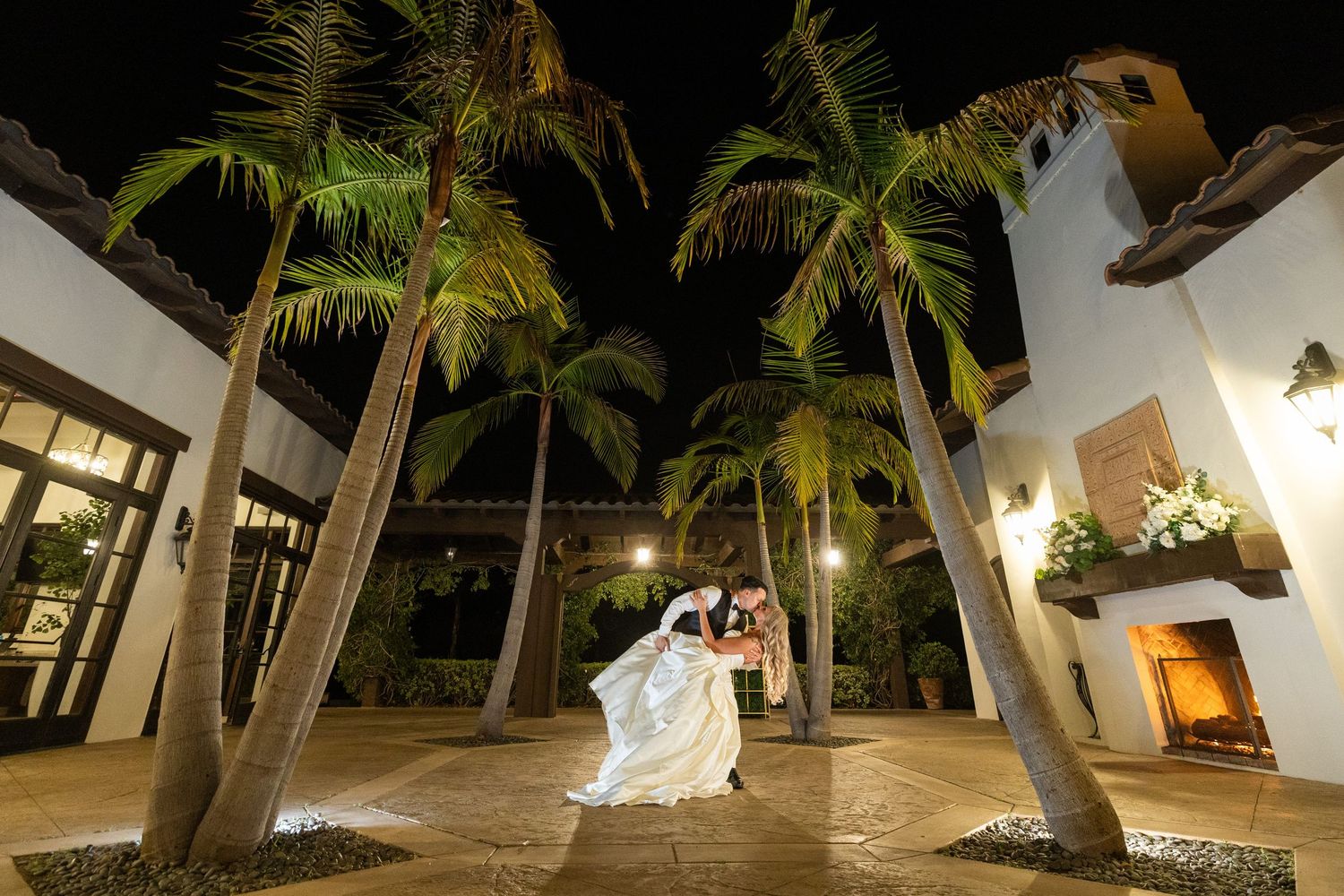 A bride in a flowing white gown with a long veil holds a bouquet of white flowers against a Mediterranean-style building.