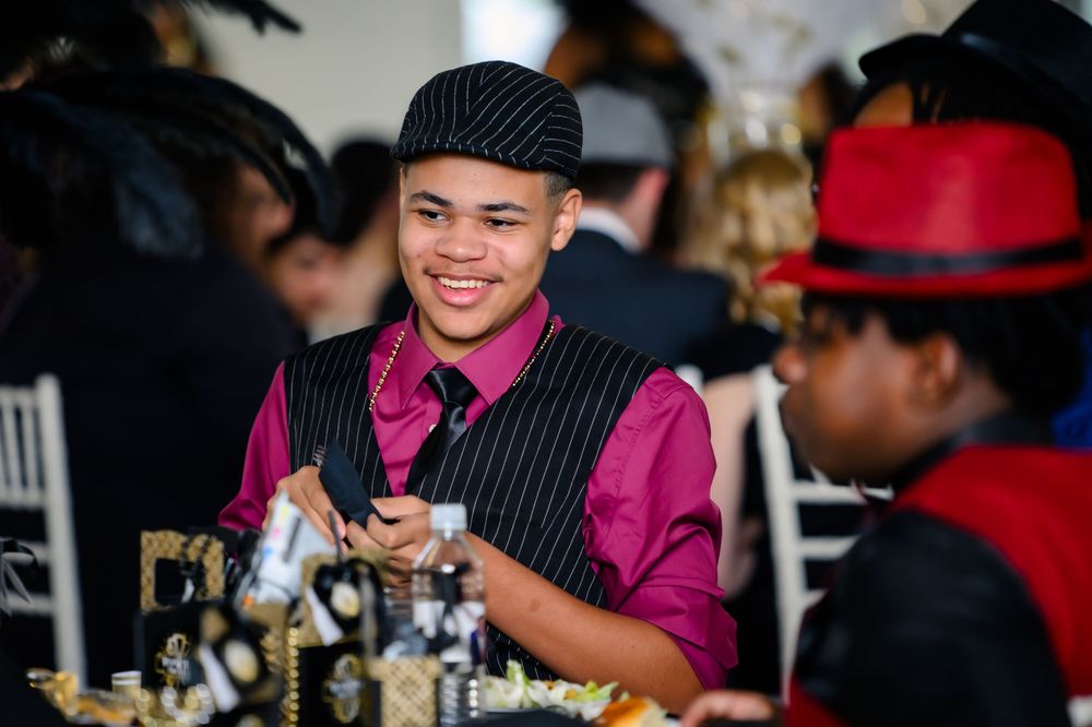 A server in a pinstriped vest and magenta shirt smiles while working at an elegant dining event.