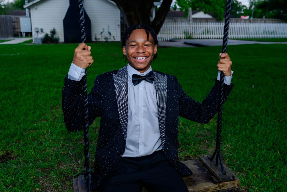 Someone wearing a black tuxedo with bowtie sits on a swing in a green backyard and smiles joyfully.