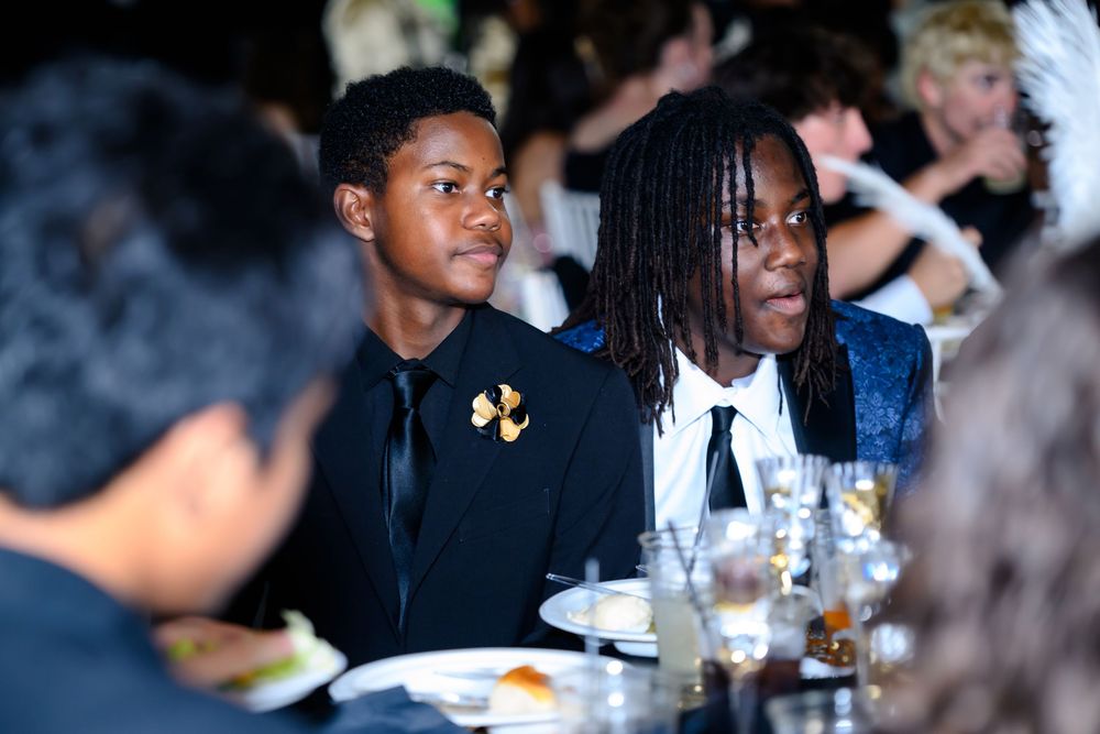 Two people seated at a formal dinner event wearing dark attire with table settings and glassware visible.