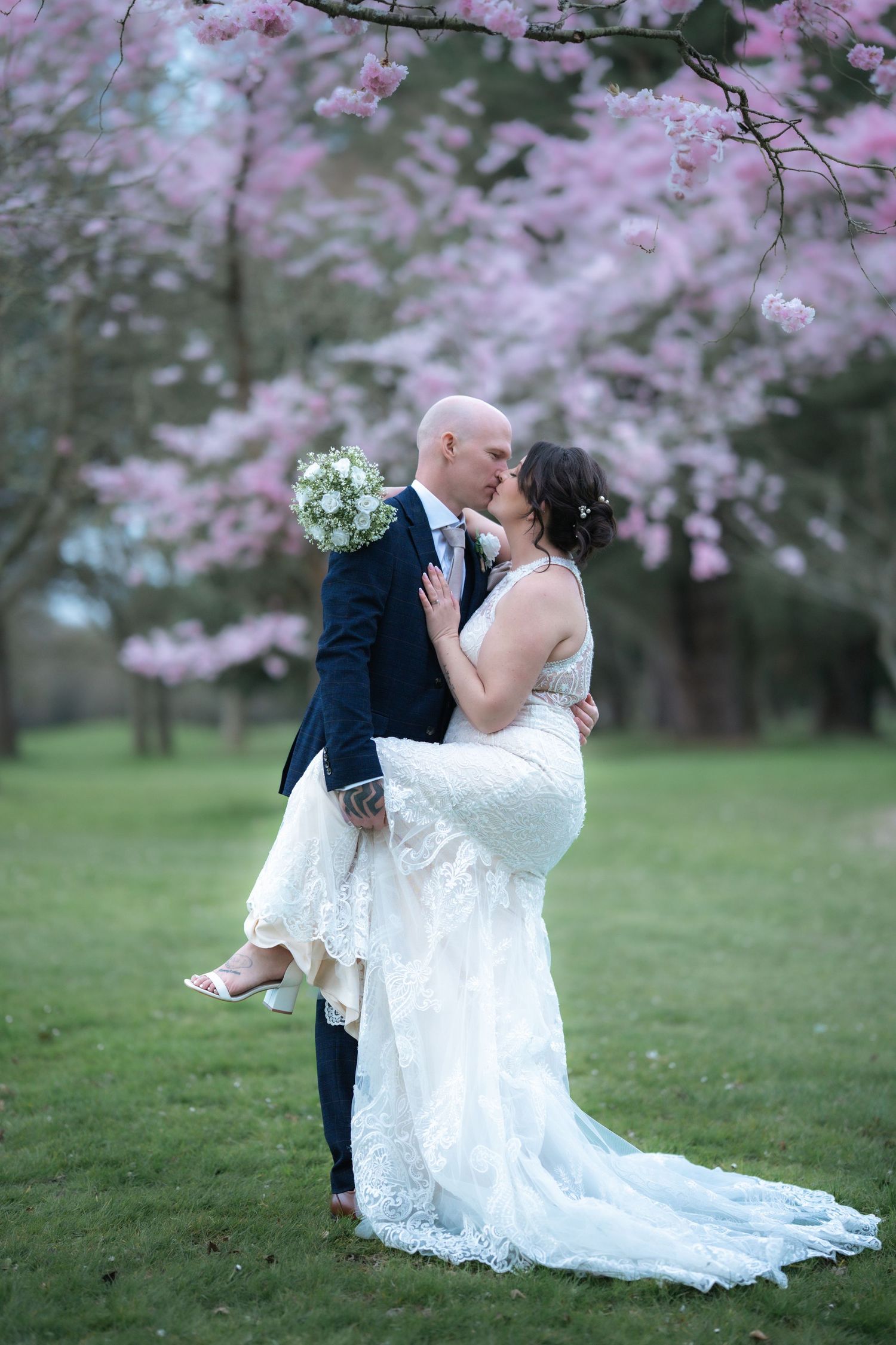 A romantic wedding photo under blooming cherry blossom trees with a white lace gown and blue suit.